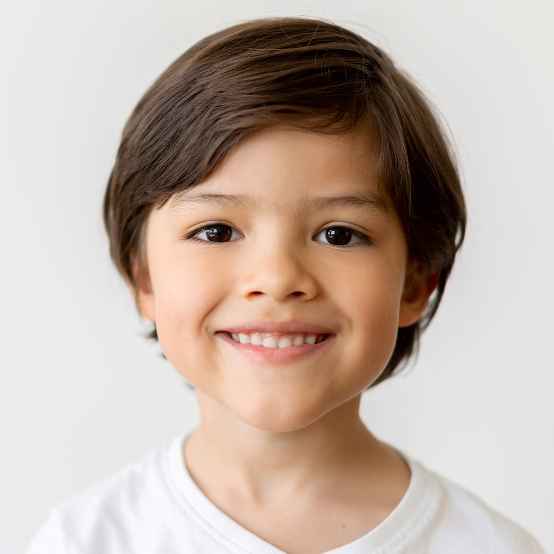 Ein kleiner junge mit braunen Haaren und braunen Augen in einem weißen T-Shirt schaut direkt in die Kamera und lächelt.